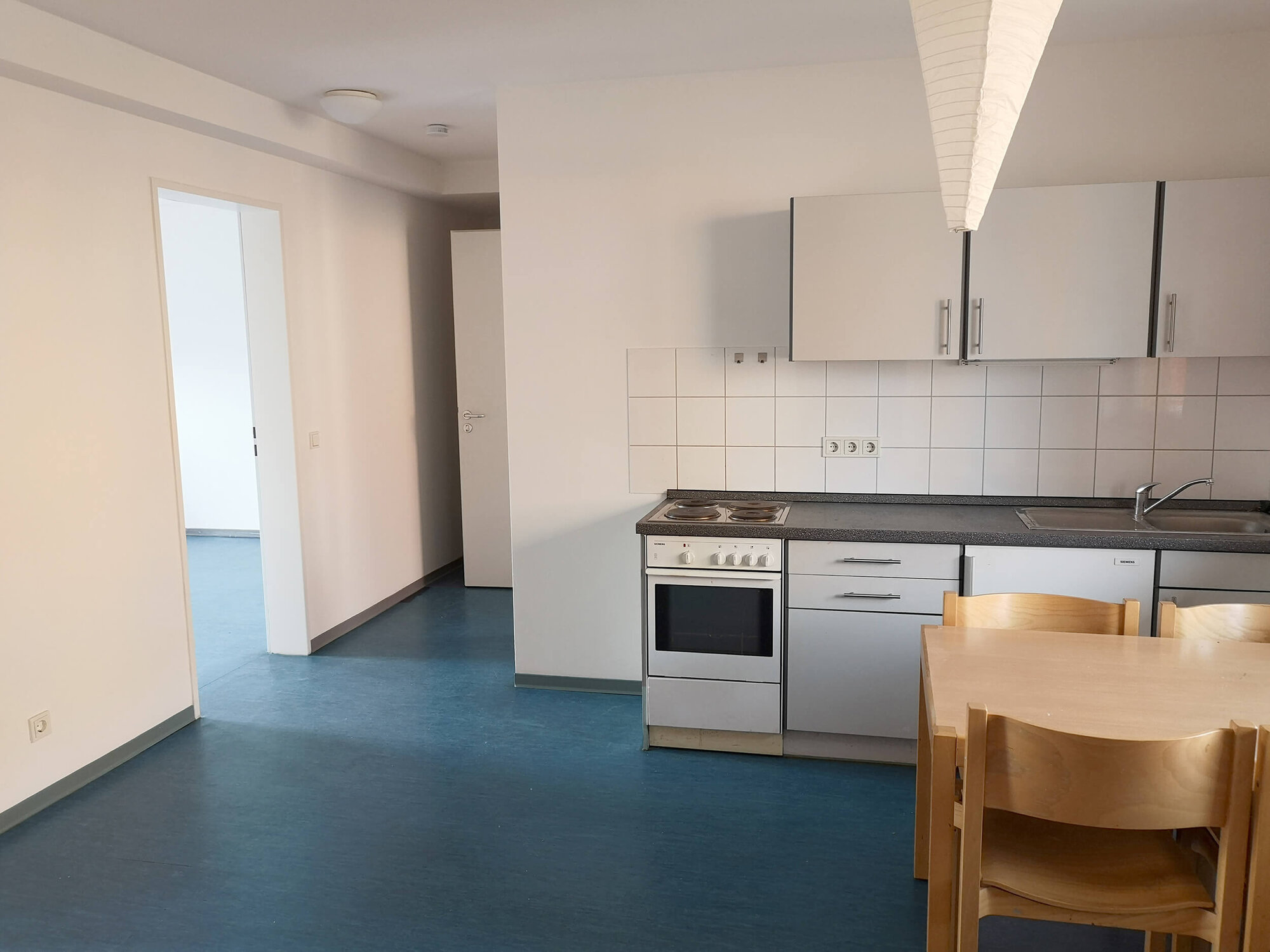 Küchenansicht mit Herd, Spüle und Esstisch in modernem, hellen Raum mit blauen Bodenbelag.