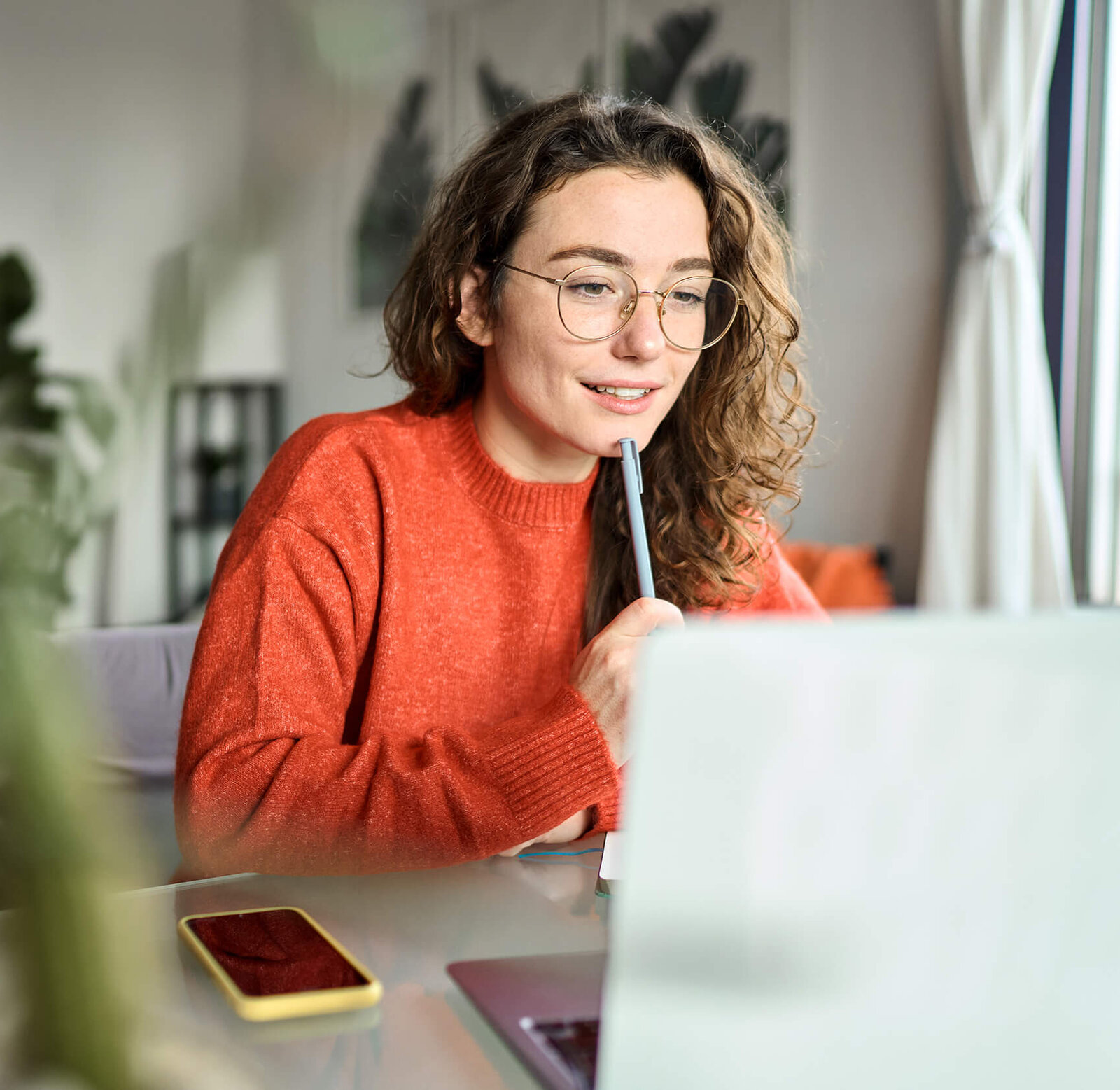 Frau mit orangenem Pullover hält Stift ans Kinn und schaut auf Laptop.