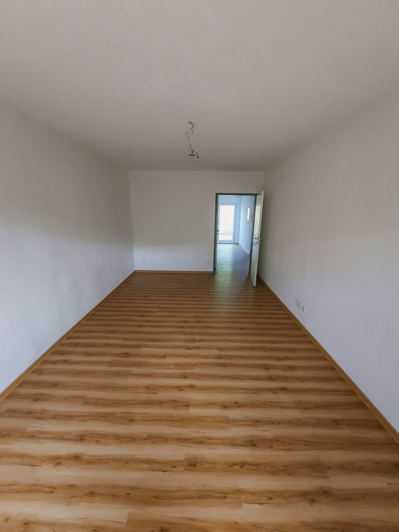 Helle und leere Wohnung mit Holzboden, Blick zur offenen Tür und viel natürlichem Licht.