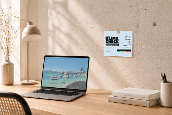Schreibtisch mit aufgeklapptem Laptop, Lampe und an der Wand befestigter Infokarte mit dem Titel 'KLIMA CHECK'.