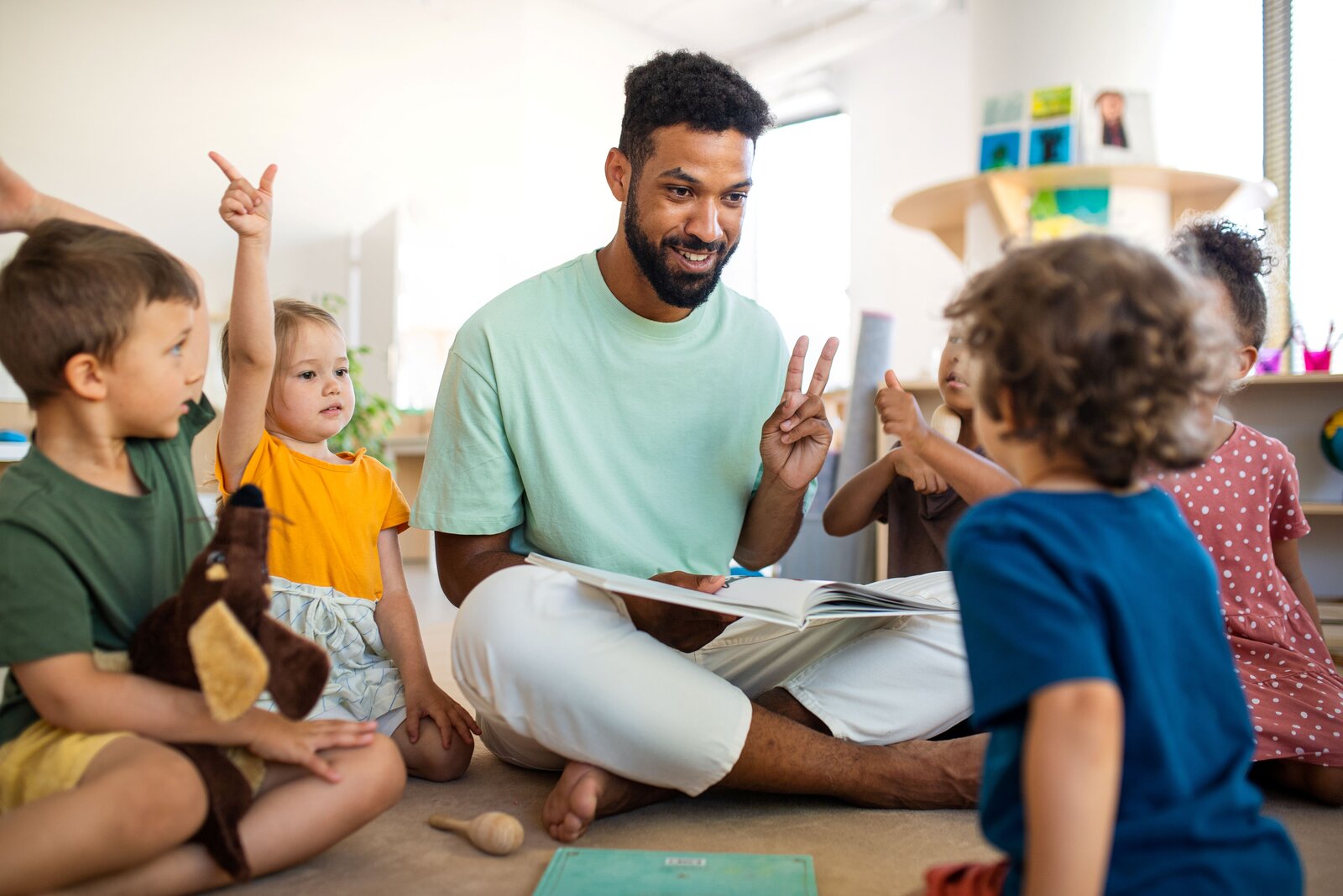 Mann mit hellblauem Shirt sitzt auf dem Boden, um ihn herum sind mehrere verschiedene Kinder.
