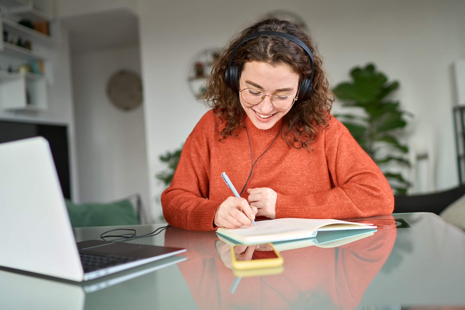 Ein lächelnder junge Frau sitzt am Schreibtisch mit Laptop, Notizbuch und Stift.