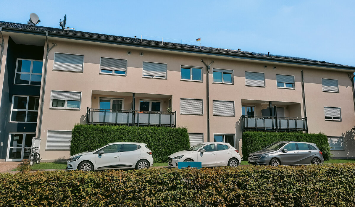 Moderne Wohnanlage im Weisenseeweg mit weißen Autos, grüner Hecke und Balkonen unter blauem Himmel.