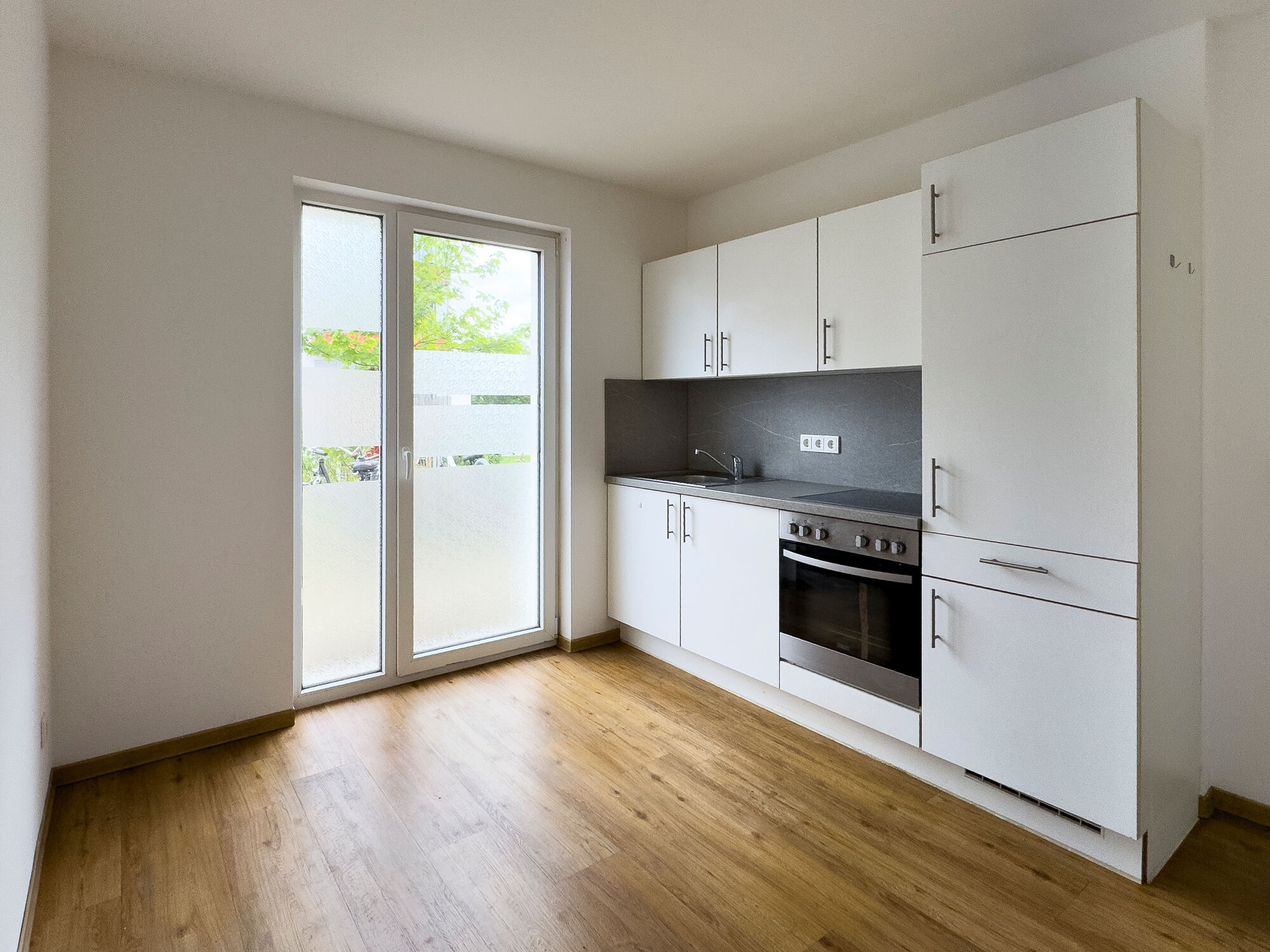 Küche mit weißen Schränken, Spüle, Elektroherd und Fenster mit Blick ins Grüne.
