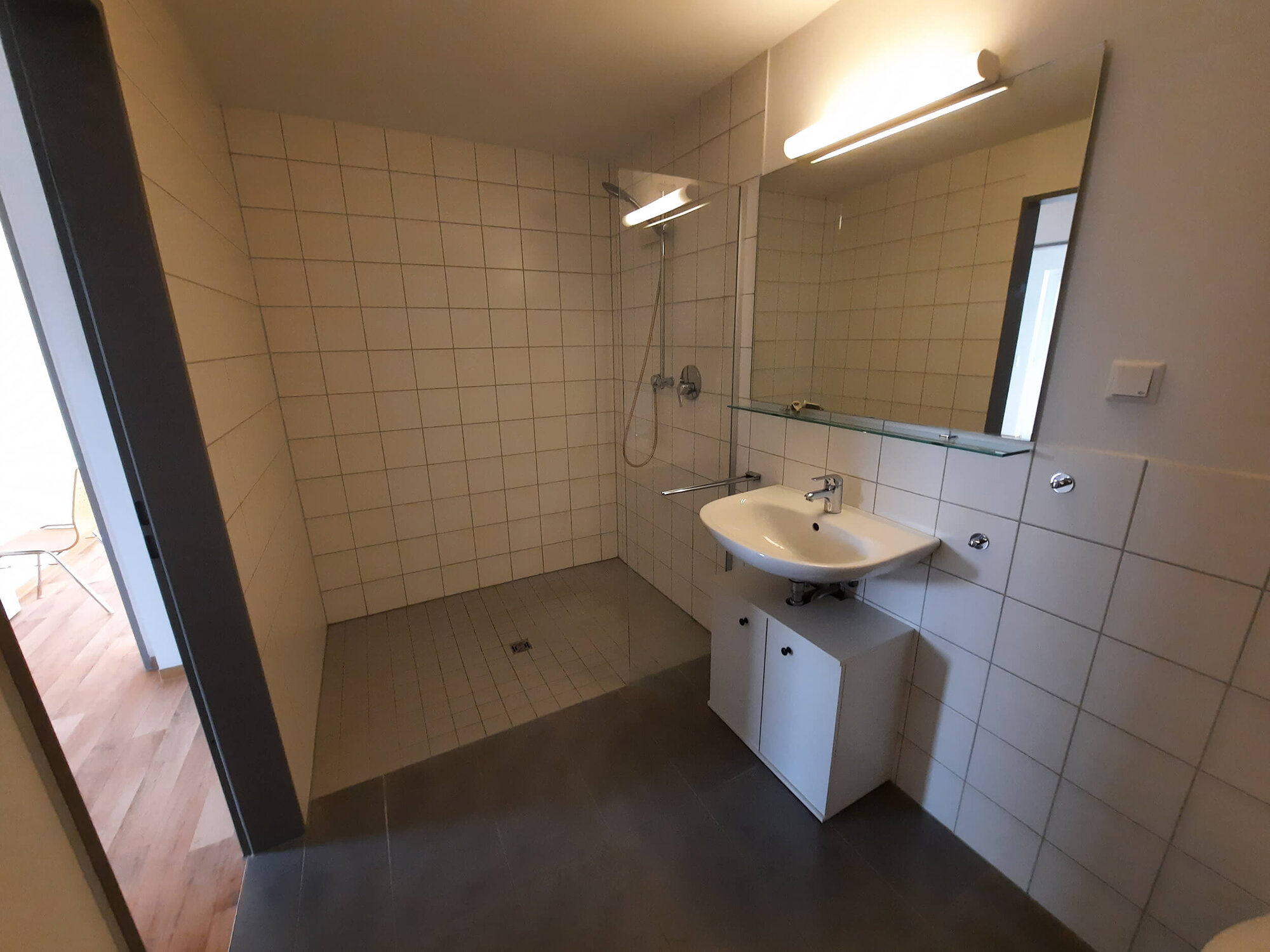 Helle Badezimmeransicht mit Dusche, Waschbecken und Stühlen im Hintergrund.