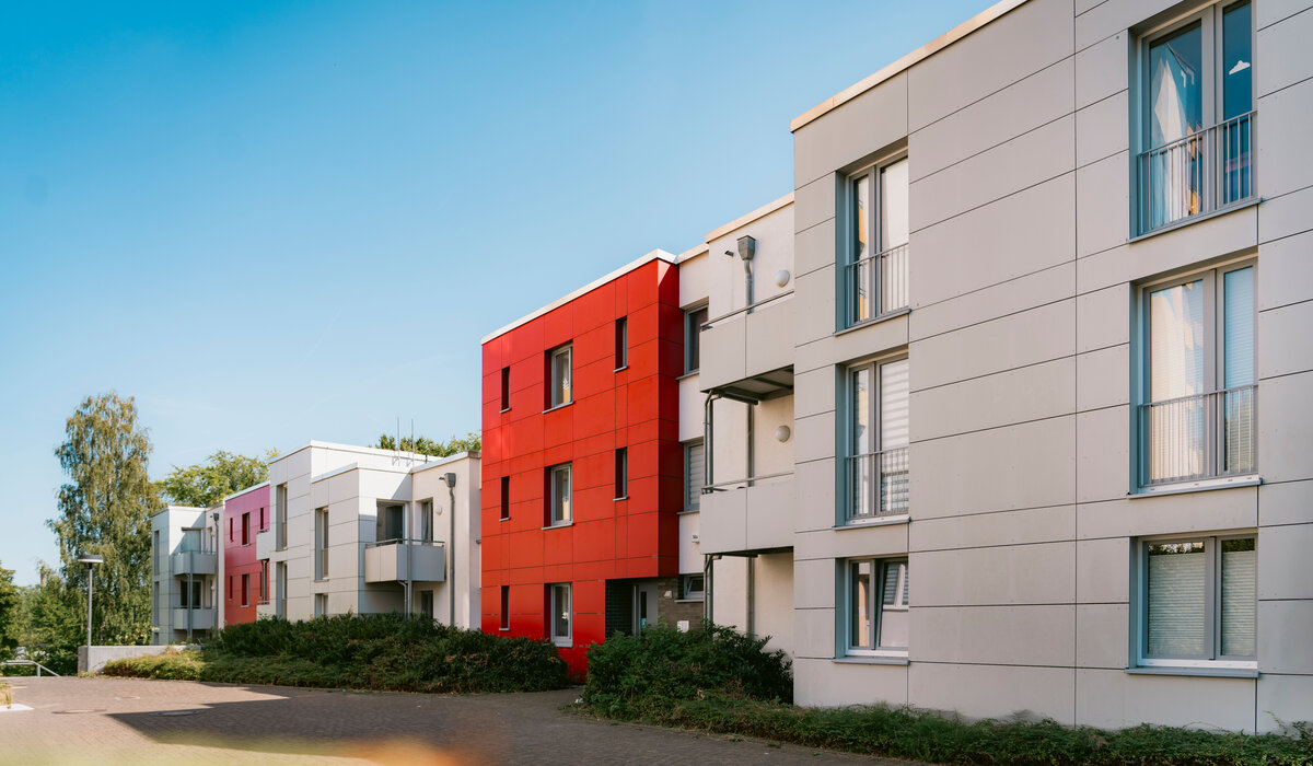Modernes Gebäude "Jakob-Kaiser-Straße 14, 14a, 14b" mit roter und weißer Fassade sowie Balkonen und grüner Bepflanzung im Vordergrund.