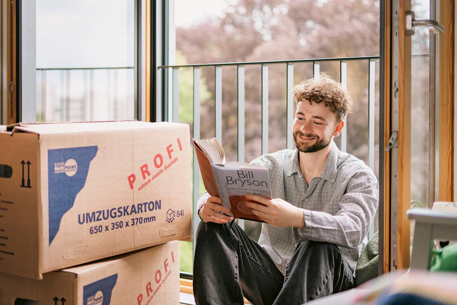 Eine Person sitzt mit aufgeschlagenem Buch neben mehreren Umzugskartons an einem Fenster mit Blick ins Freie.