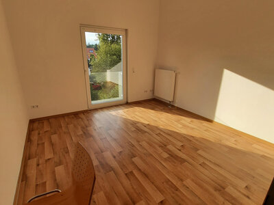 Helles Zimmer mit Holzfußboden, großer Fenstertür und einem Stuhl in der unteren Ecke.