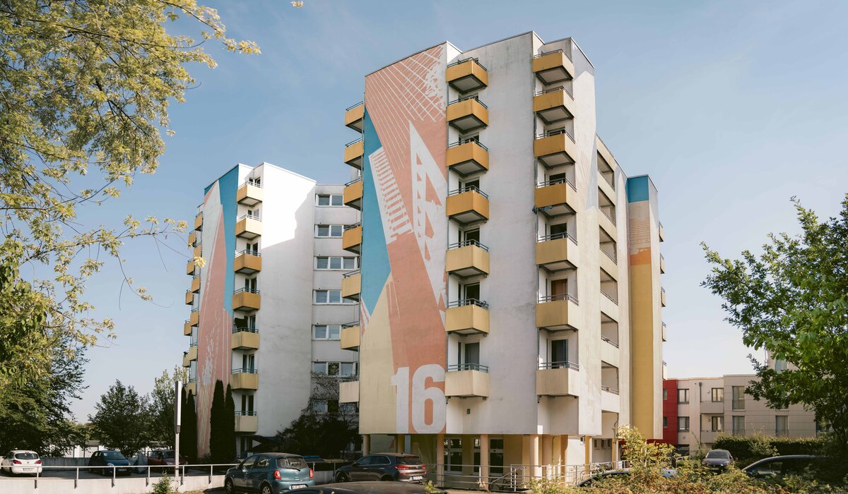 Wohnanlage "Jakob-Kaiser-Straße 16" mit buntem Fassadenkunstwerk, Balkonen und blühendem Baum im Vordergrund.