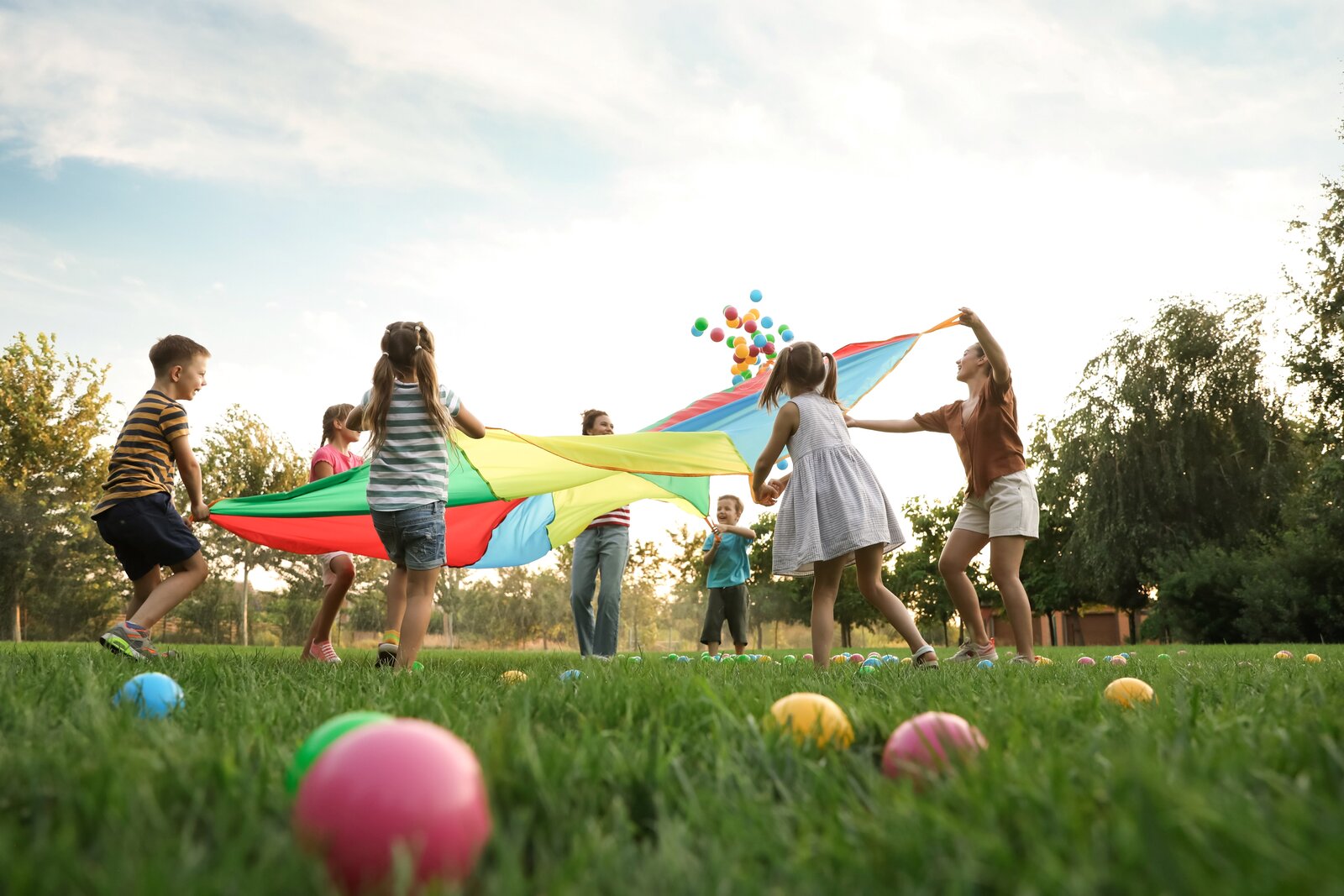 Kinder spielen im Freien auf einer Wiese mit einem bunten Schwungtuch; mehrere bunte Bälle fliegen in die Luft, weitere liegen im Gras, während die Sonne hinter Bäumen im Hintergrund scheint.
