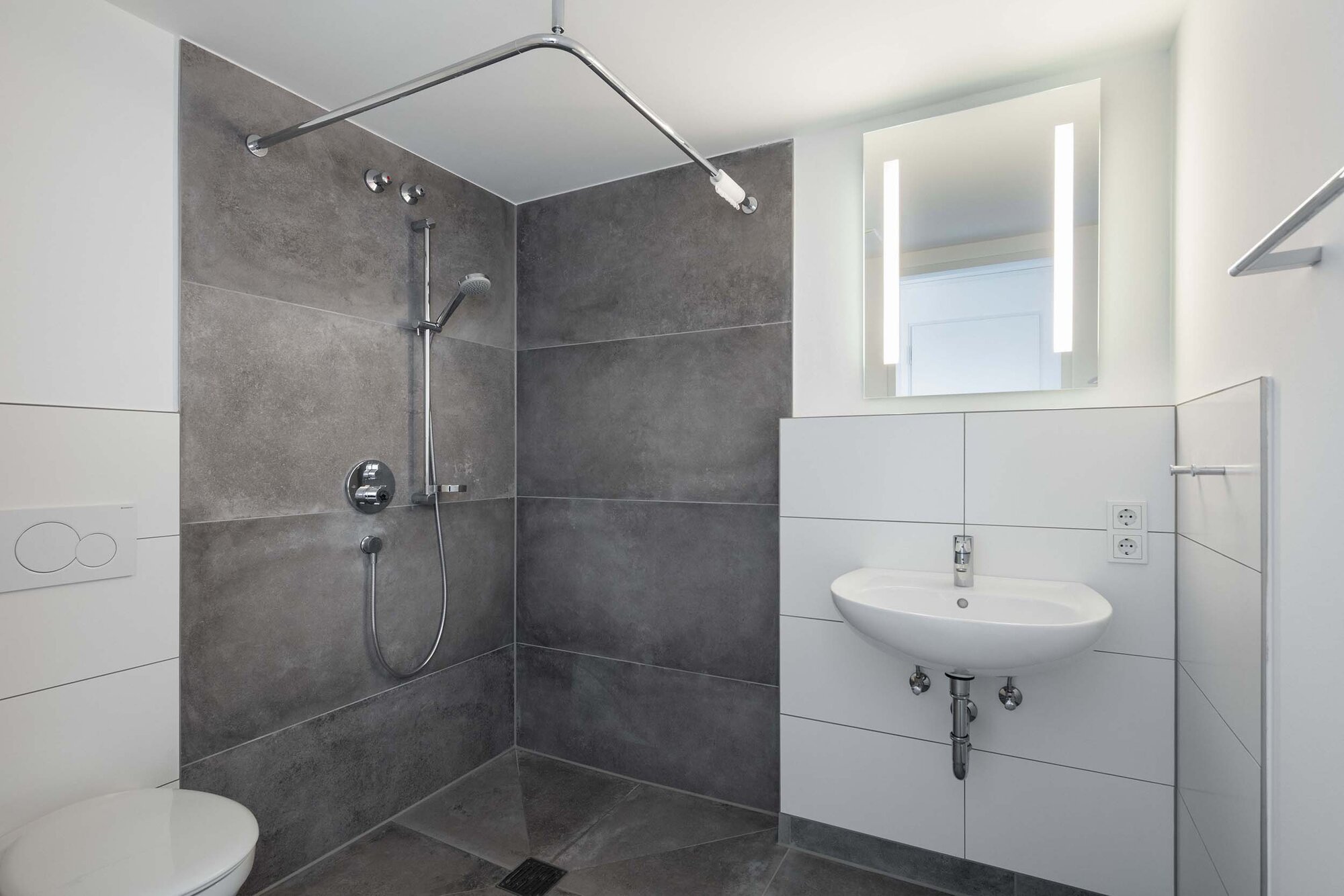 Moderne Dusche mit grauen Fliesen, Handbrause, Waschbecken und LED-Spiegelbeleuchtung.