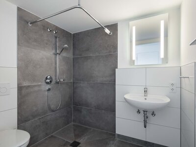 Moderne Dusche mit grauen Fliesen, Handbrause, Waschbecken und LED-Spiegelbeleuchtung.