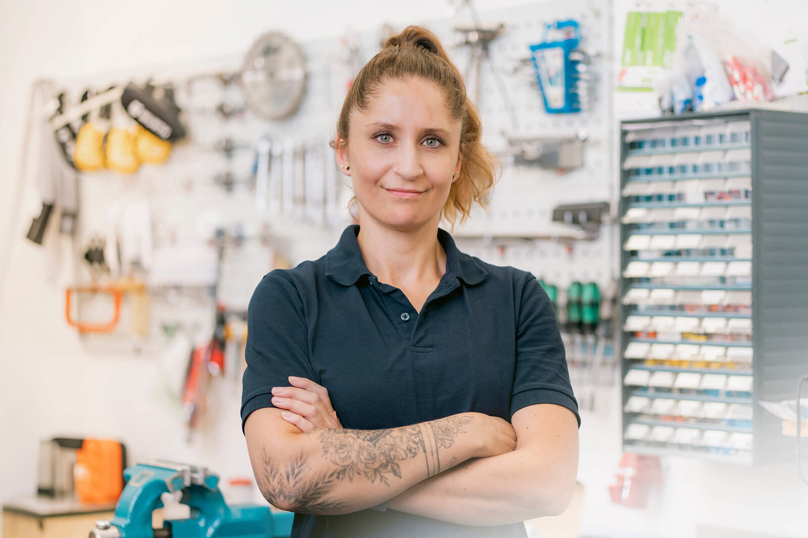 Junge Frau mit Poloshirt und verschränkten Armen, im Hintergrund hängen verschiedene Werkzeuge an der Wand.