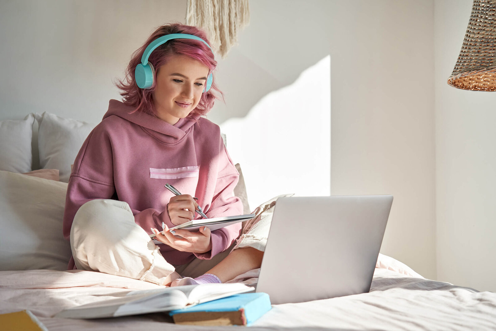 Eine junge Frau mit Kopfhörern sitzt auf einem Bett und schreibt in ein Notizbuch vor einem aufgeklappten Laptop.
