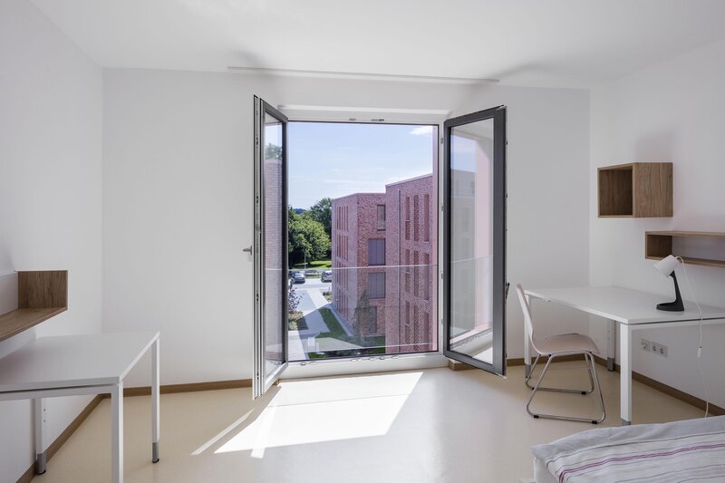 Helles Zimmer mit offener Fenstertür, Tisch, Stuhl und Ausblick auf den Außenbereich.