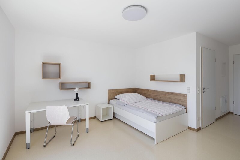 Schlafzimmer mit schmalem Bett, Schreibtisch, Stuhl und Wandregalen in minimalistischer Einrichtung.