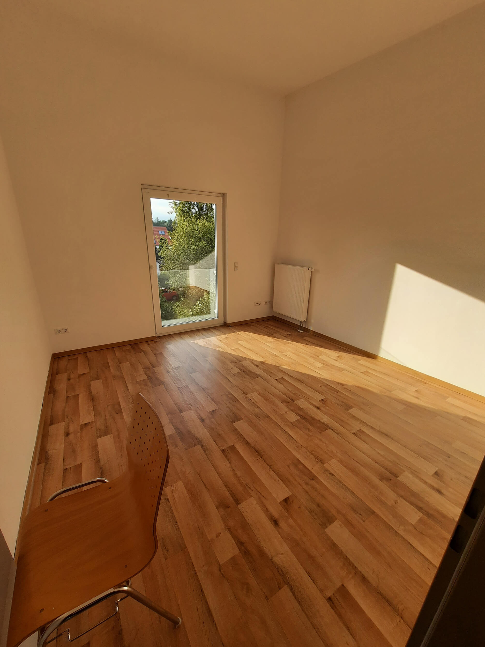 Helles Zimmer mit Holzfußboden, großer Fenstertür und einem Stuhl in der unteren Ecke.