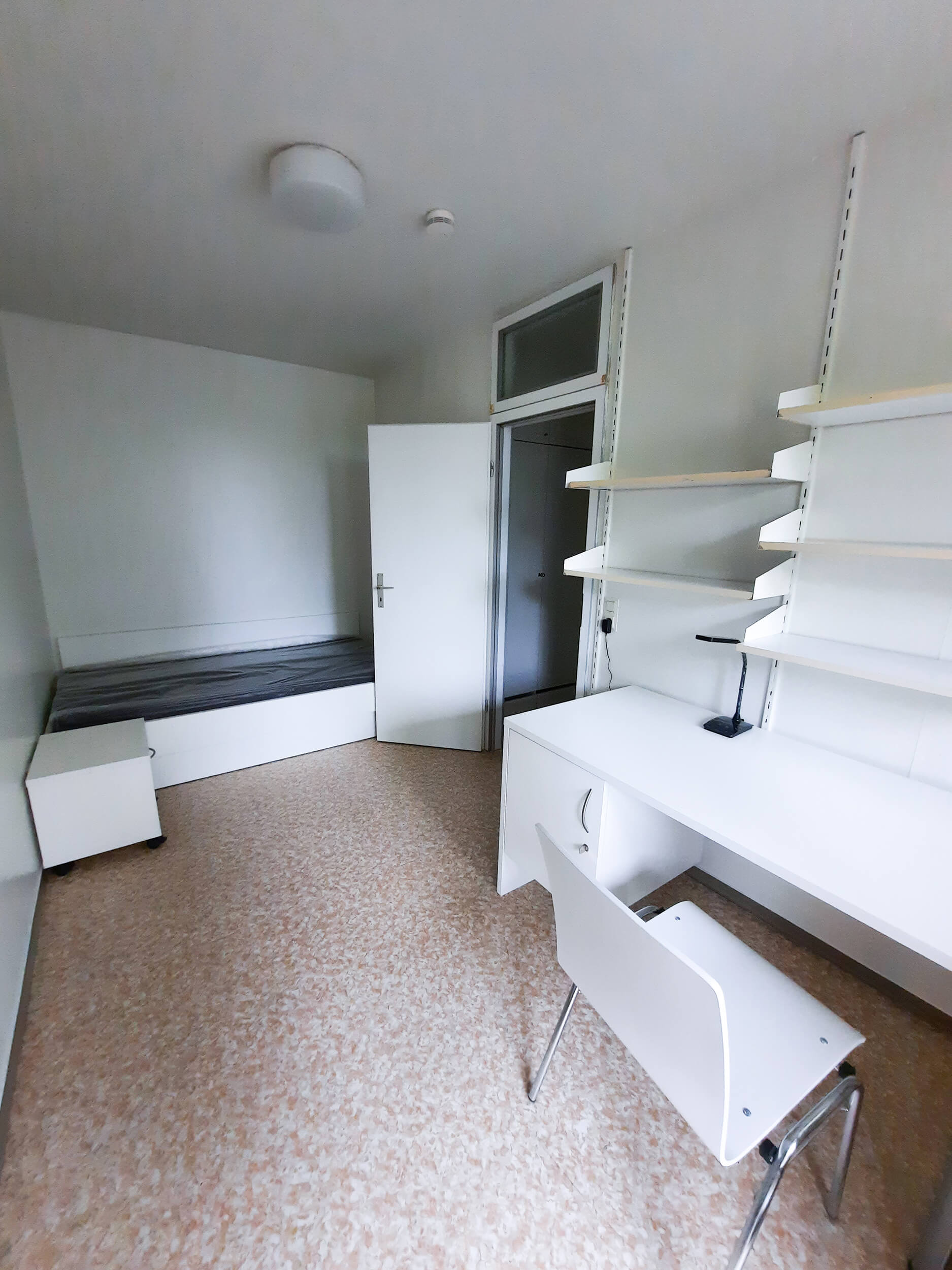 Helles Zimmer mit Bett, Schreibtisch, Stuhl und Regalen für Stauraum.