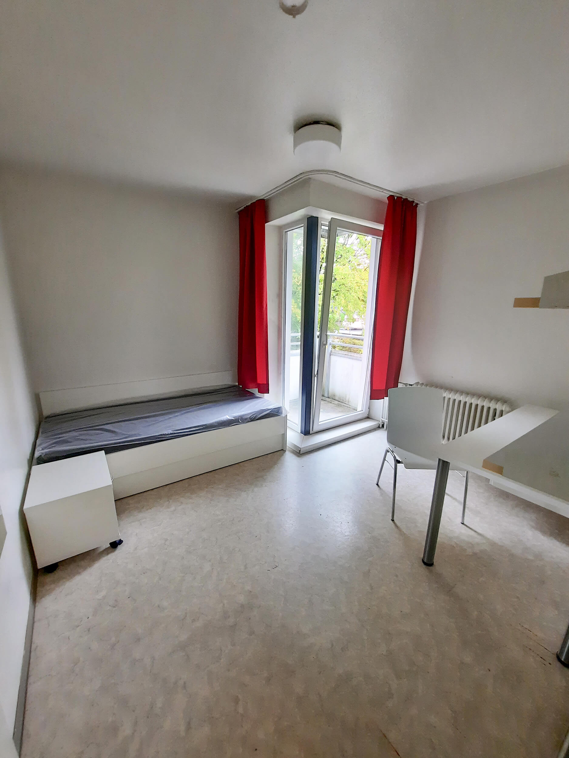 Helle Studiozimmer mit rotem Vorhang, Einzelbett, Tisch und Stuhl sowie Zugang zum Balkon.